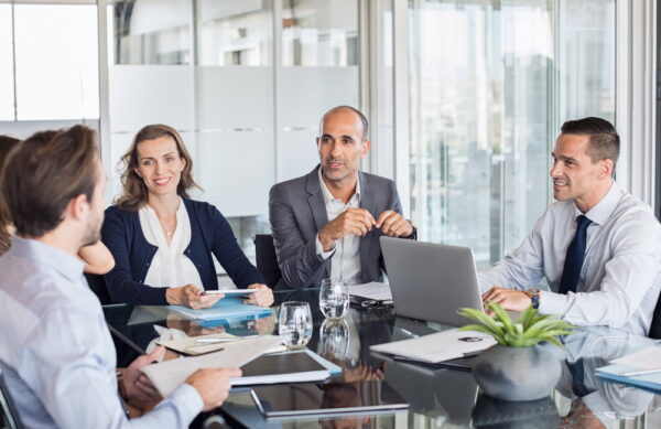 Multiethnic group of business people sitting in annual meeting in conference room. Mature leader with businessmen and businesswomen discussing. Busy partners working together in modern office.